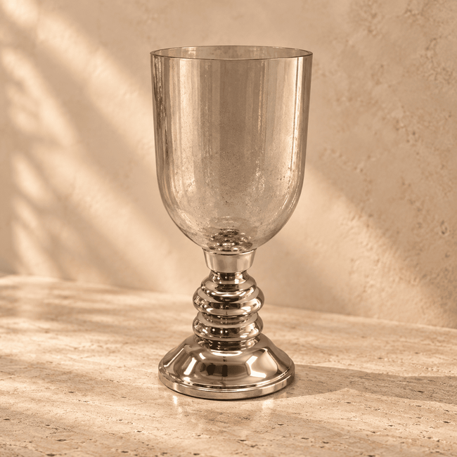 A clear glass goblet with a polished silver-toned pedestal base, set on a textured beige stone surface against a matching beige wall with soft shadow patterns.