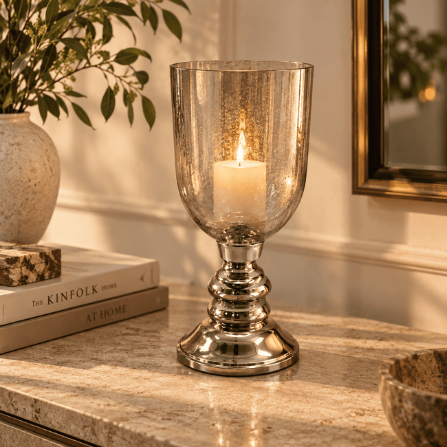 A clear seeded glass candle holder with a polished silver-toned pedestal base, holding a lit white candle, set on a marble countertop in a warm, sunlit interior.